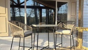 Une terrasse couverte avec deux fauteuils en osier et une table, de grandes fenêtres et une vue sur les arbres.