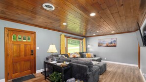 Uma sala de estar com teto de madeira, uma janela grande, um conjunto de sofás e um quadro na parede.