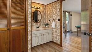 Um banheiro com porta de madeira, um lavatório com espelho e uma mesa de bilhar verde ao fundo.