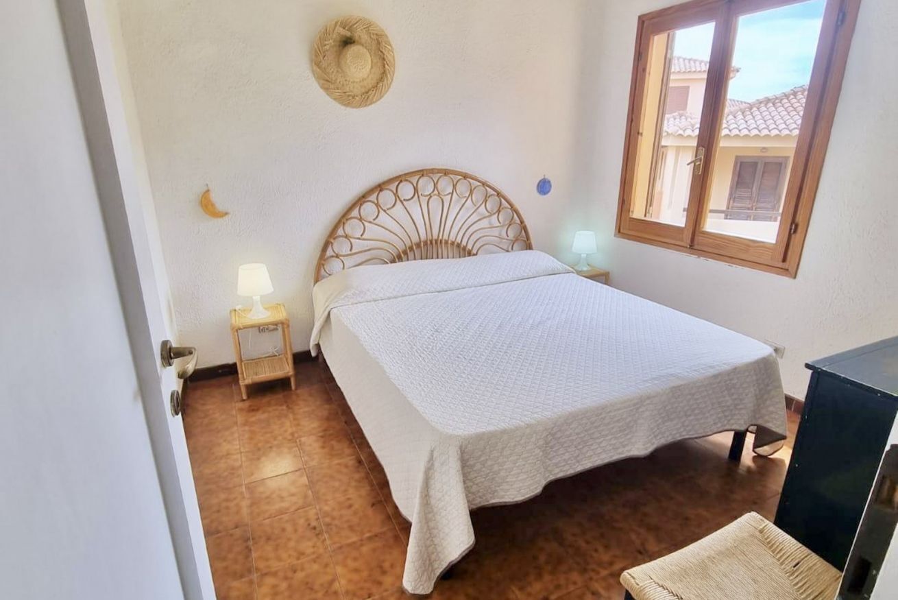 Ein Schlafzimmer mit Bett, Nachttisch, Wandhaken für Hüte, einem Fenster und einer Tür.