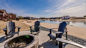 Une terrasse au bord du lac, dotée de chaises Adirondack et d’un foyer, offrant une vue sur un lac paisible et des immeubles résidentiels.