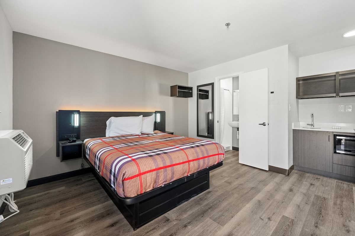 A modern hotel room with a large bed, a bedside table, a built-in heater, a small desk, and a kitchenette.