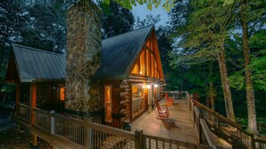 A log cabin with a stone chimney, a wooden deck, and a porch with red chairs. - Modern Log Cabin with all the Amenities (Todd)