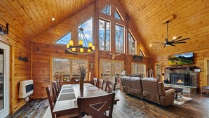 A spacious log cabin interior with a dining area, a fireplace, and a large window overlooking the outdoors. - Modern Log Cabin with all the Amenities (Todd)