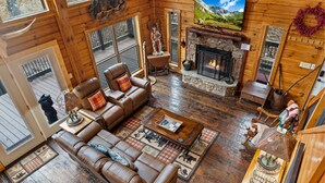A wooden cabin interior with a fireplace, leather sofa, and a large window overlooking a deck. - Modern Log Cabin with all the Amenities (Todd)