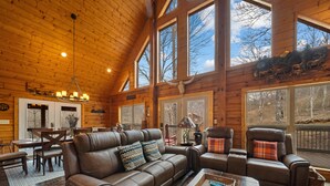 A spacious living room with a large brown leather sofa, a wooden coffee table, and a fireplace. There are large windows allowing natural light and views of the outdoors. - Modern Log Cabin with all the Amenities (Todd)