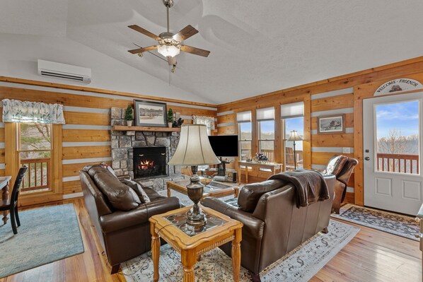 L’intérieur chaleureux d’un chalet avec une cheminée en pierre, des canapés en cuir, une table basse en bois et un ventilateur de plafond.