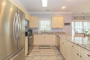 A kitchen with a stainless steel refrigerator, wooden cabinets, a granite countertop, and a window with blinds.