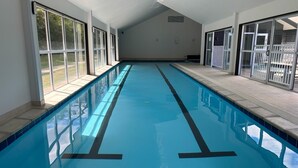 En lang, indendørs swimmingpool med krystalklart, blåt vand, omgivet af store vinduer og flisegulv.
