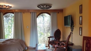 A bedroom with a bed, a wooden table, a chair, a TV, and a window with curtains.