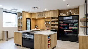 A modern, well-lit store with wooden accents and a central island. It features a refrigerated section labeled "COLD" and shelves stocked with various products. - Tru By Hilton Kalamazoo (Kalamazoo)