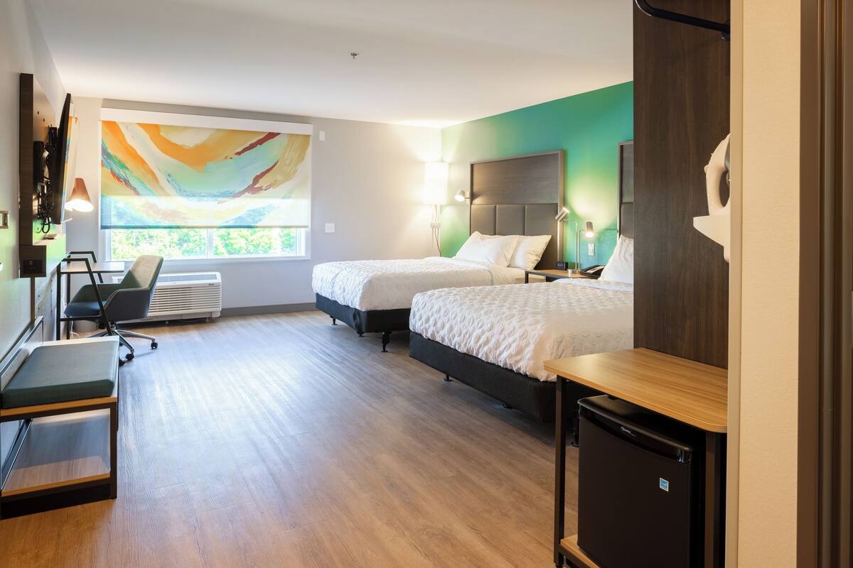 A hotel room with two beds, a desk, a chair, a small refrigerator, and a large window with a view of greenery.