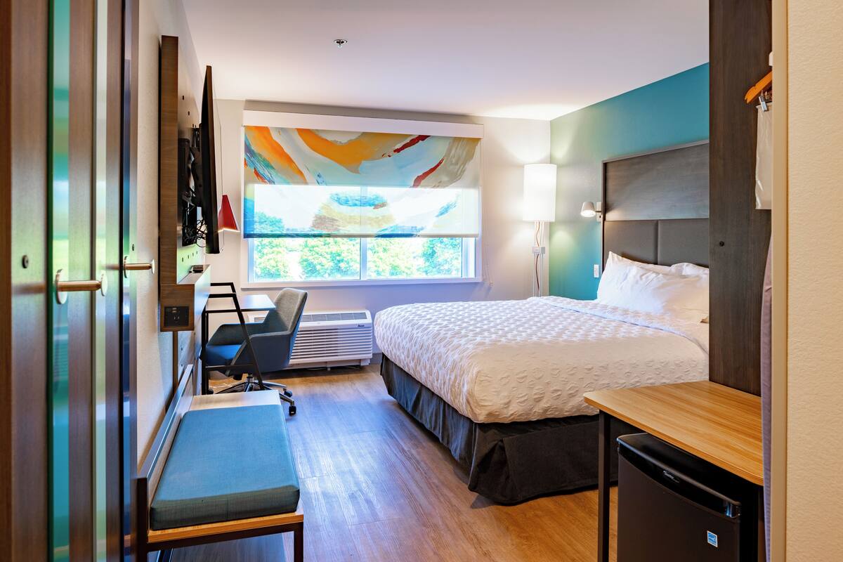 A modern hotel room with a bed, desk, chair, and a large window with a view.
