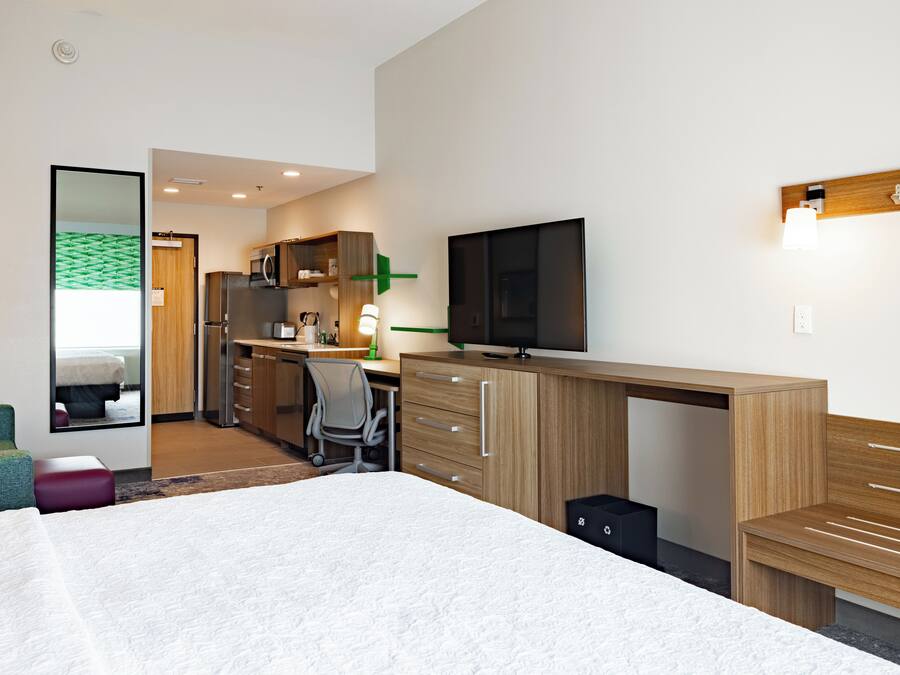 A modern hotel room with a large bed, a desk with a computer, a television, and a small kitchenette.