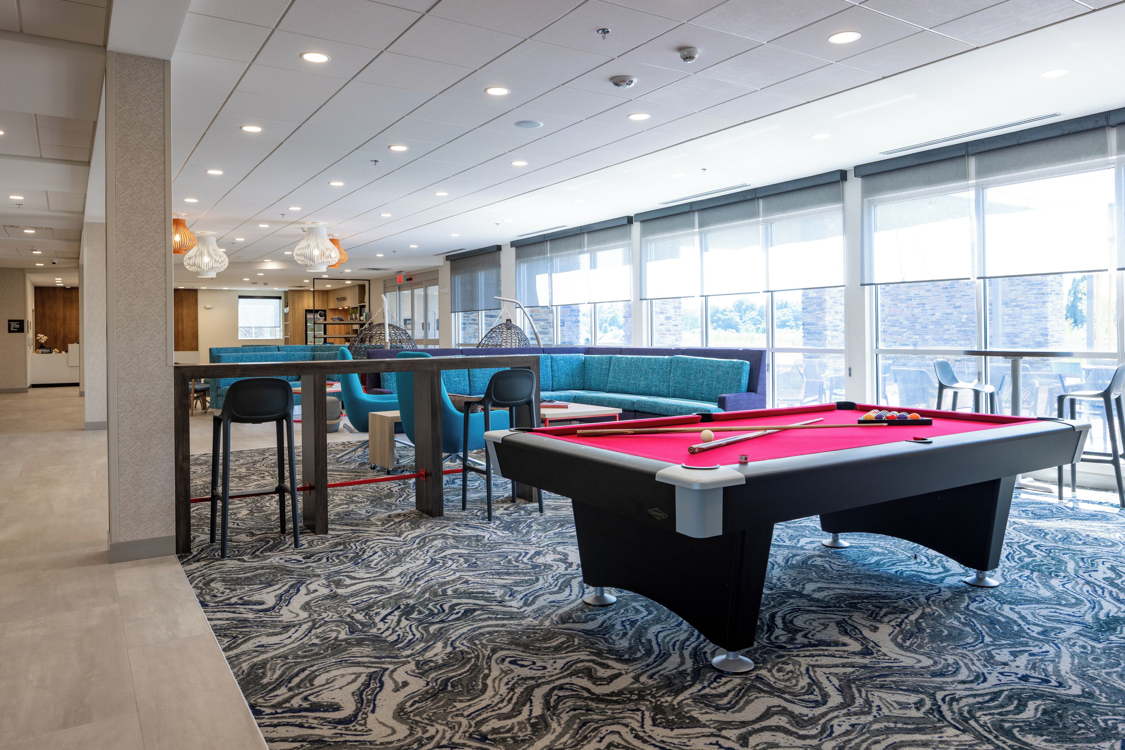 A pool table in a modern room with a blue sofa and chairs, large windows, and a bar area.