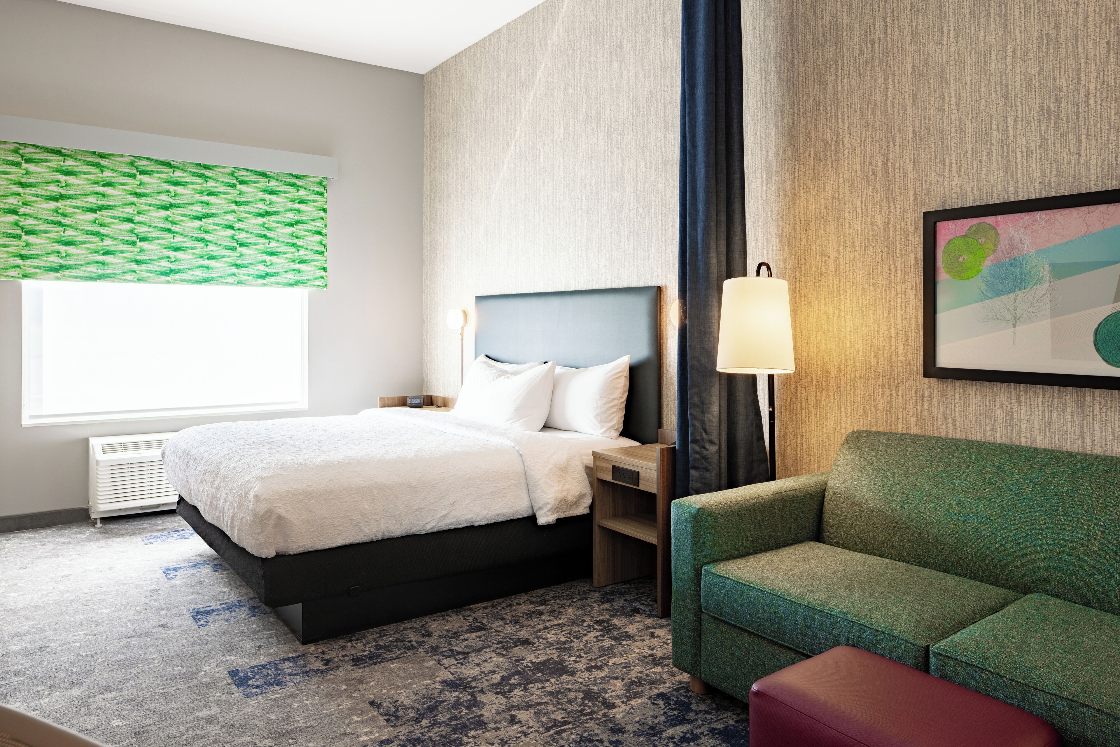 Habitación de hotel con cama, sofá verde, mesita de noche, lámpara y un cuadro en la pared.