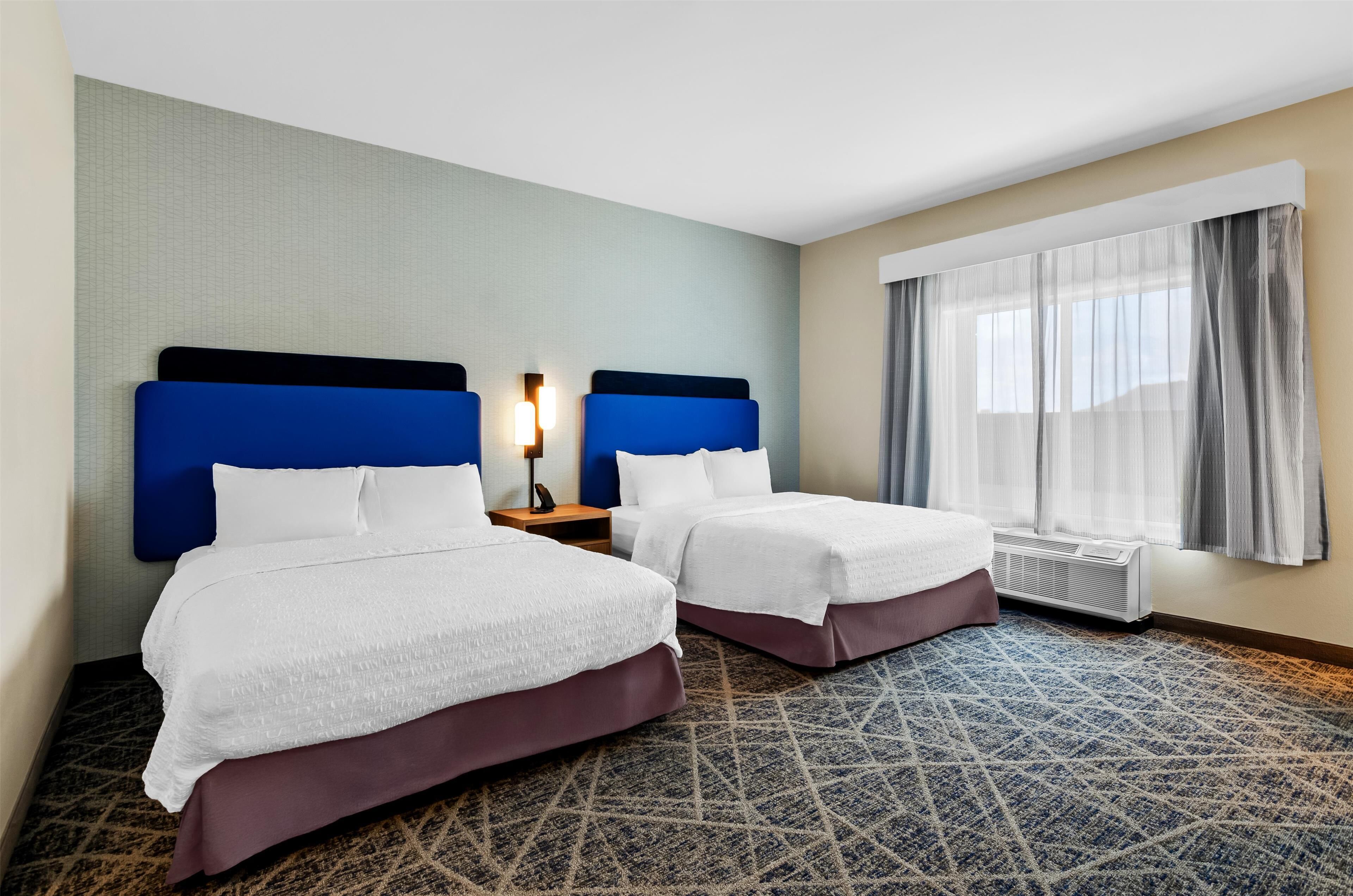 A hotel room with two beds, a patterned carpet, a window with curtains, and a wall-mounted heater.