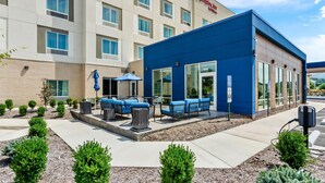 A modern hotel with a blue exterior, outdoor seating area with blue chairs and umbrellas, and a landscaped garden with small trees. - Hampton Inn Daleville (Daleville)