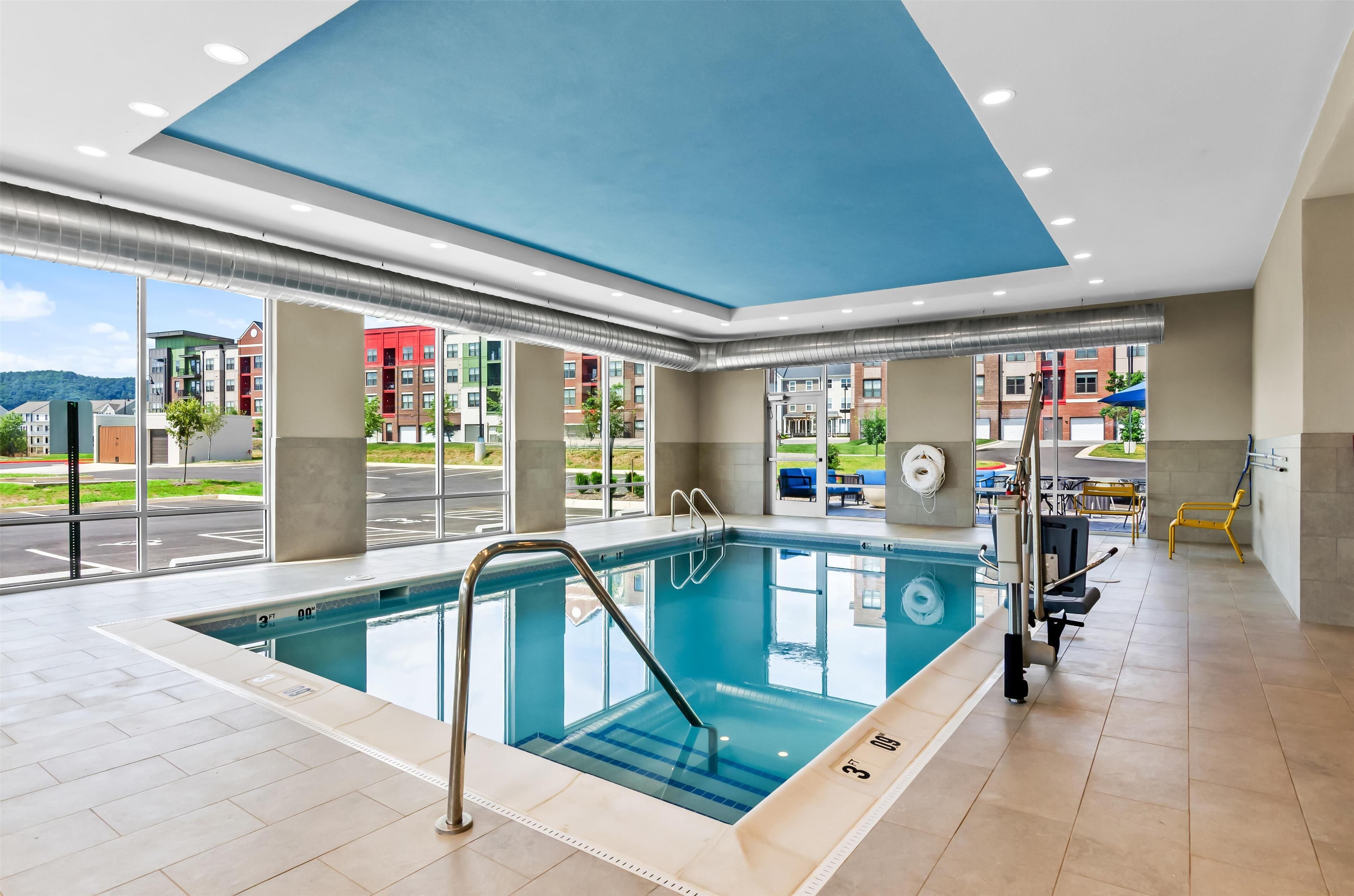 Una piscina interior moderna con paredes de vidrio transparente, techo azul y vistas al exterior.