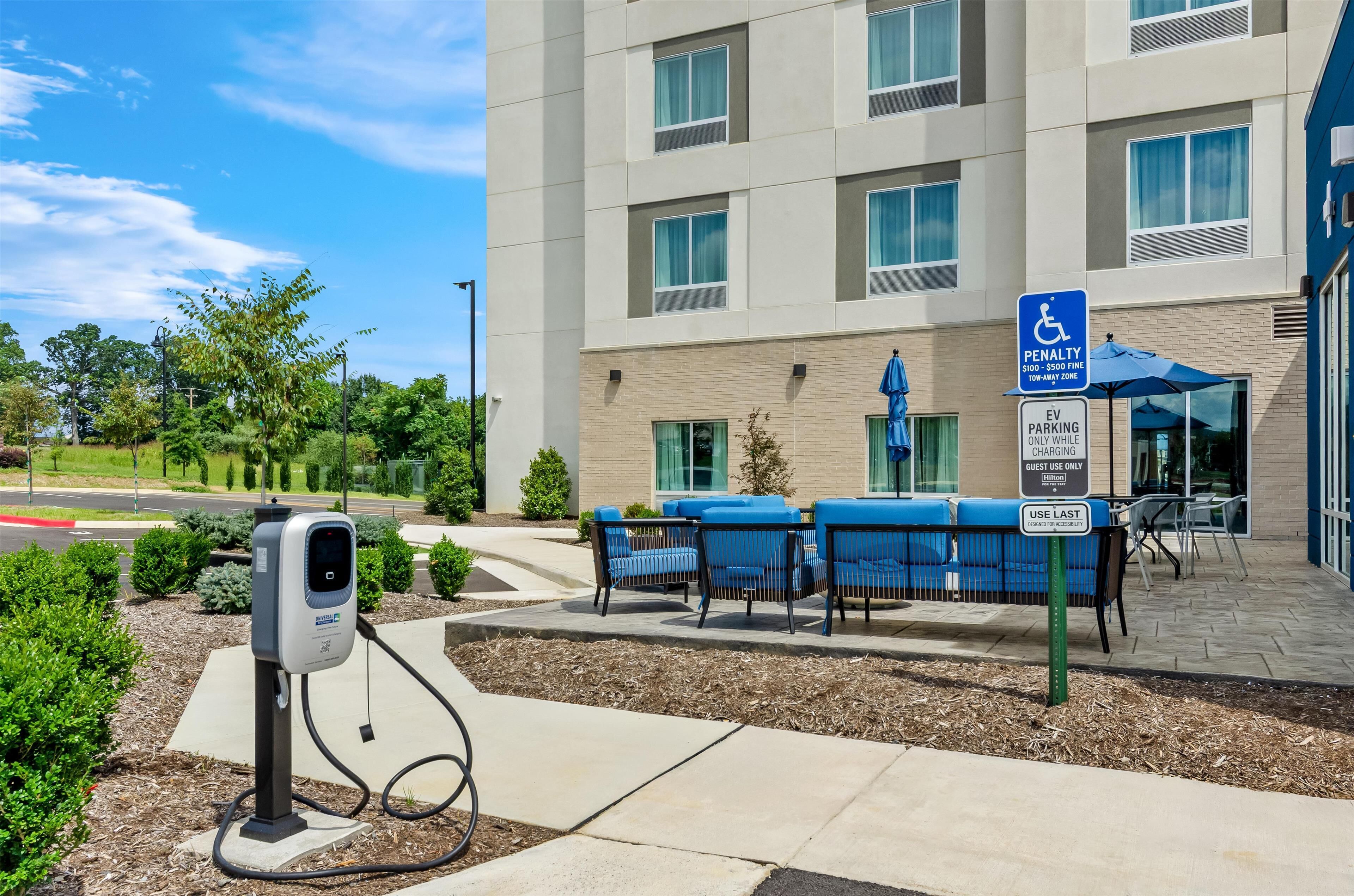 Un estacionamiento con estaciones de carga para vehículos eléctricos, un edificio con varias ventanas y mobiliario exterior con sillas azules.