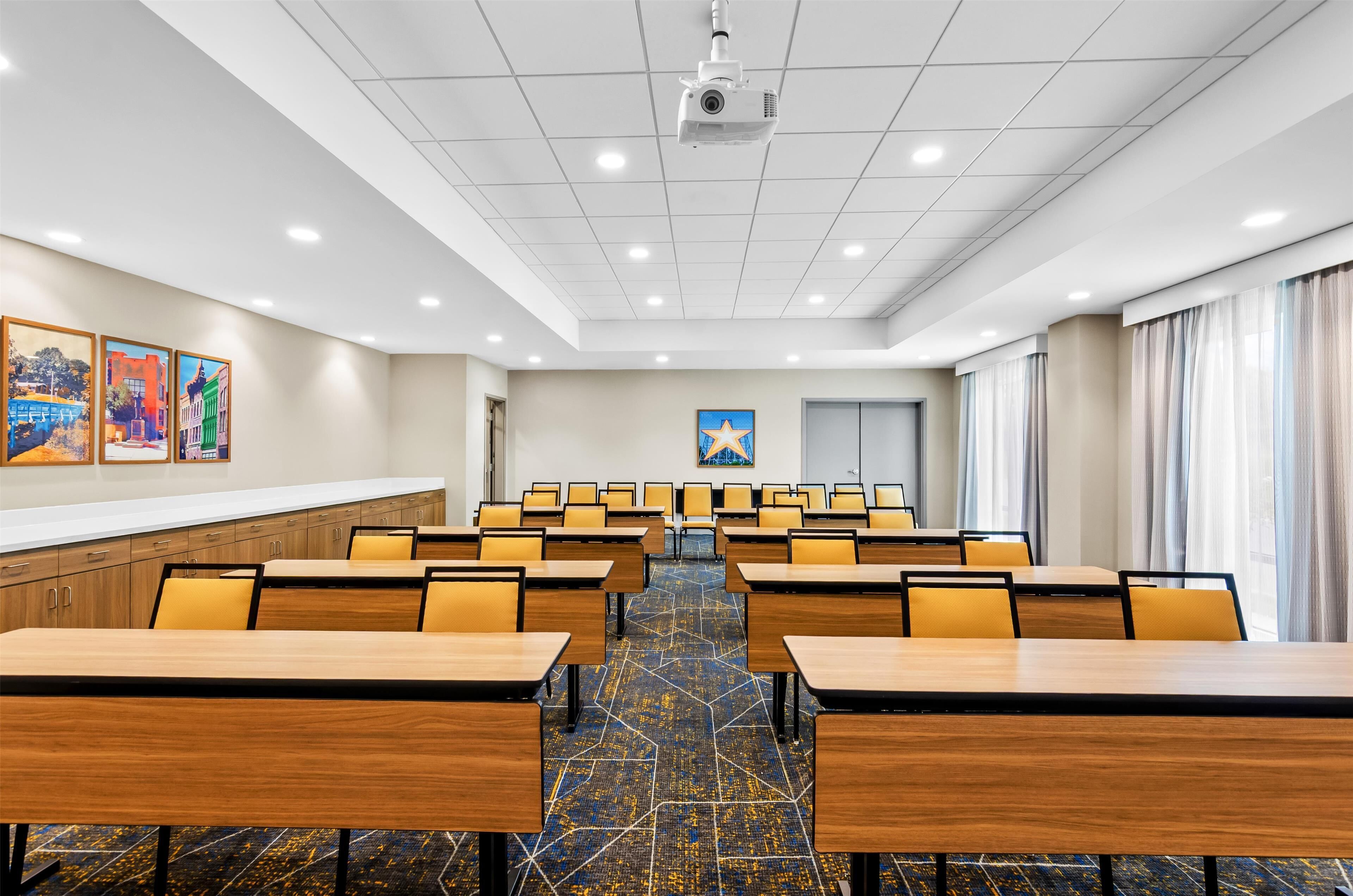 Una sala conferenze con file di tavoli in legno e sedie gialle, un proiettore a soffitto e una decorazione a forma di stella sul muro.