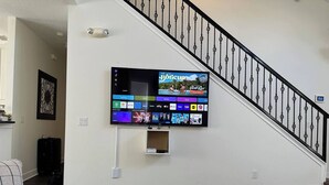 Una sala de estar con un televisor de pantalla plana montado en la pared, una escalera con barandilla negra y un sofá gris.
