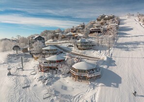 Estância de esqui coberta de neve com vários edifícios, incluindo um grande hotel de montanha e chalés menores, ligados por um teleférico.