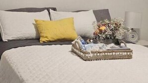 Un letto rifatto con copriletto bianco trapuntato, due cuscini (uno giallo), un cesto di vimini con un'infiorescenza e un dipinto floreale sulla parete.
