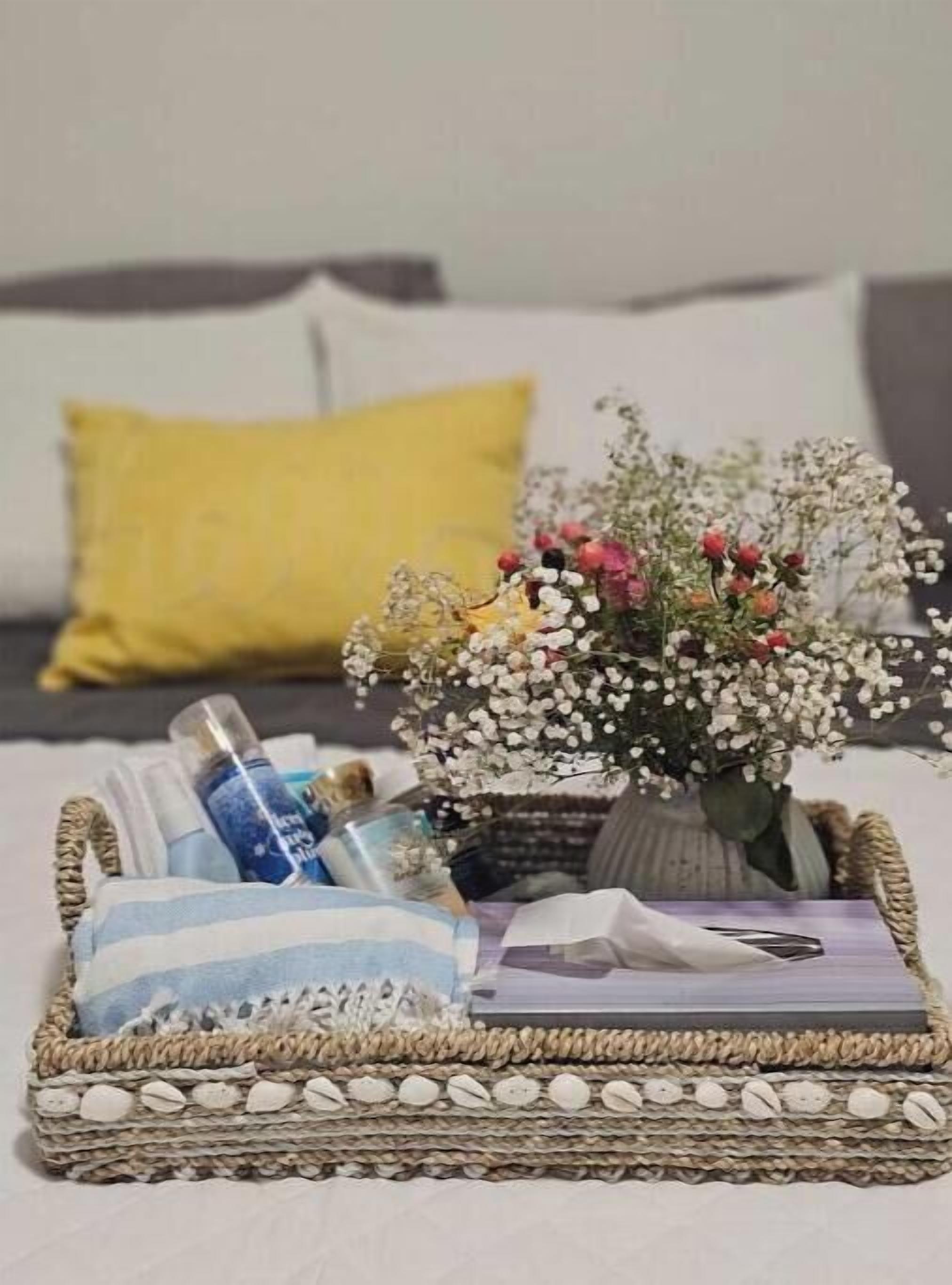 Un plateau en osier avec un vase de fleurs, une bouteille et quelques livres, posés sur un lit agrémenté d’un coussin jaune.