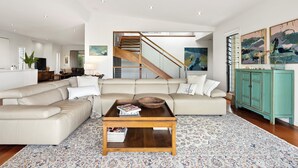 Un salon spacieux avec un grand canapé en forme de L, une table basse en bois et un tapis à motifs. Un escalier avec des rampes en bois mène à l’étage supérieur.