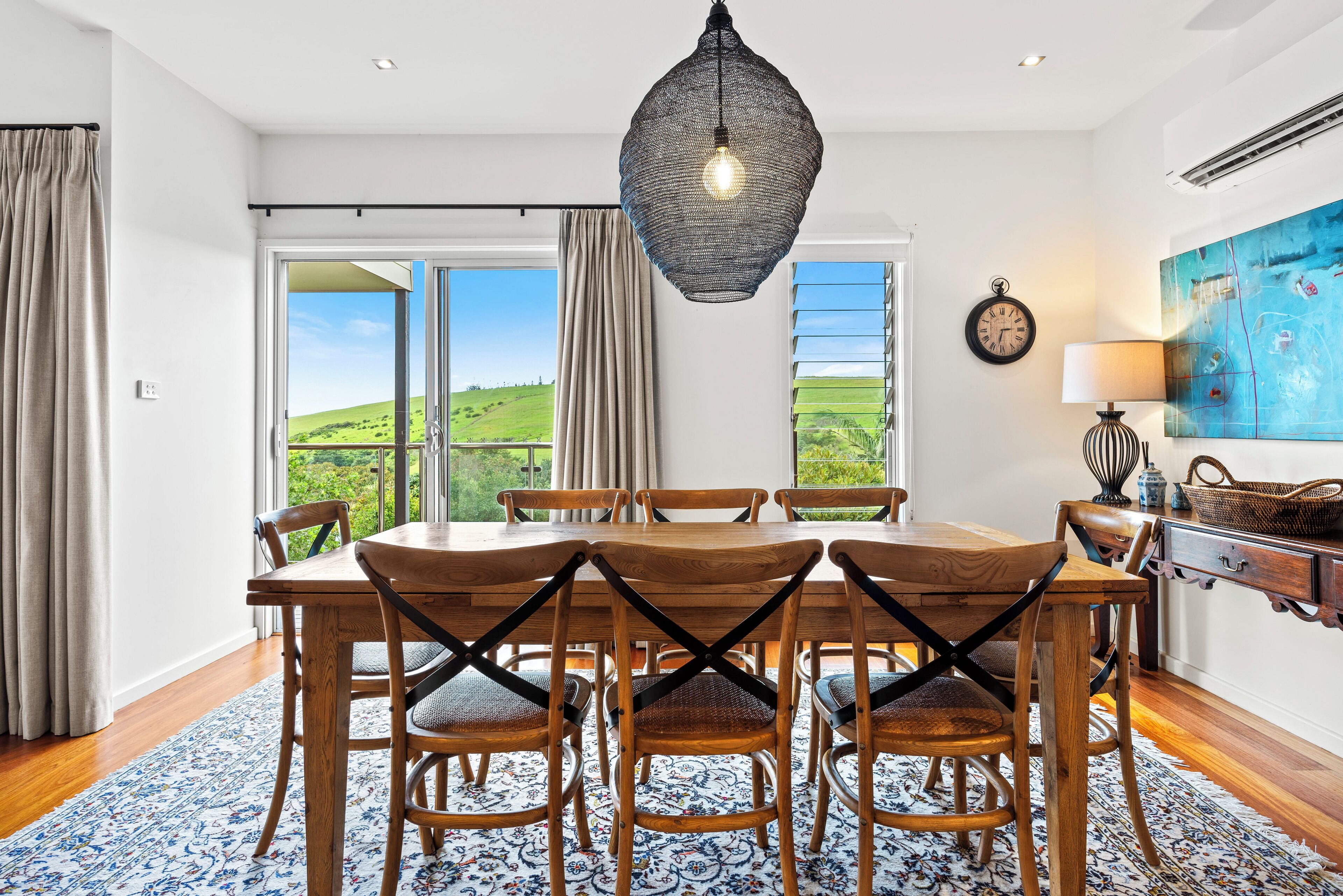 A dining area with a wooden table and chairs, a hanging light fixture, and a view of green hills through large windows.