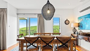 Un coin repas avec une table et des chaises en bois, un luminaire suspendu et une vue sur des collines verdoyantes à travers de grandes fenêtres.
