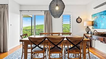 A dining area with a wooden table and chairs, a hanging light fixture, and a view of green hills through large windows.