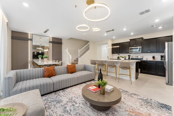 A modern living room with a grey sofa, a coffee table, and a kitchen area with dark cabinets and a stainless steel refrigerator.