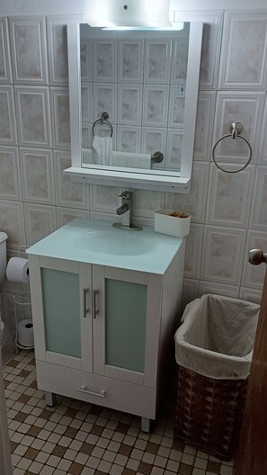 Un baño con un lavabo blanco, un espejo encima y un cesto de basura a la derecha.