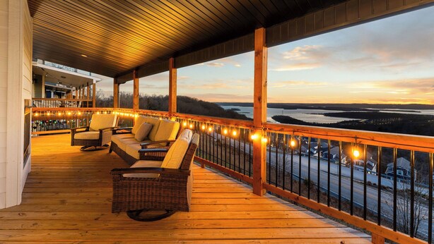 Una terraza de madera con mobiliario de mimbre y luces colgantes, con vistas a un paisaje pintoresco.