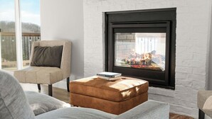 Un salón con chimenea de ladrillo blanco, un sillón color beige, un reposapiés marrón y un ventanal con vistas a una terraza.