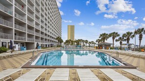 Zona de piscina con sillones de descanso, un edificio alto al fondo y palmeras.