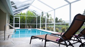 A pool area with a glass enclosure, two lounge chairs, and a view of a tropical landscape.