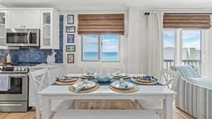 Een eetgedeelte met een witte tafel gedekt voor vier personen, een keuken met witte kastjes en een magnetron, en uitzicht op de zee door een raam met bruin zonnescherm.