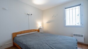 A bedroom with a wooden bed frame, a grey bedspread, a bedside lamp, a window with blinds, and a wall-mounted heater. - Apartment, terrace, beach within walking distance (Agde)