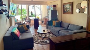 A living room with a sofa, a coffee table, and a TV. There is a ceiling fan, a painting on the wall, and a refrigerator. - Casa Corazon Unit 702 - Ocean View, huge terrace in beautiful Puerto Vallarta! (Puerto Vallarta)