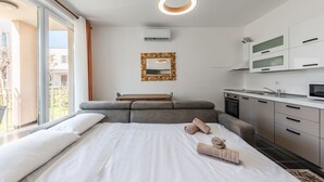 A modern hotel room with a large bed, a small sofa, a kitchen area, and a balcony with a view.
