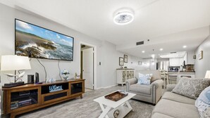 A modern living room with a flat-screen TV, a wooden entertainment unit, a grey sofa, and a kitchen in the background.