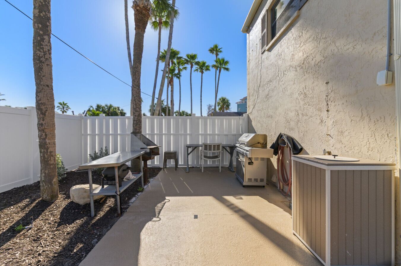 A backyard with a barbecue grill, a table, and chairs, surrounded by a white fence and palm trees.