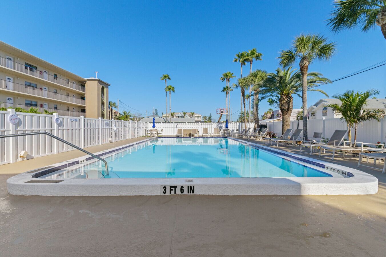 Ein Pool mit einer Tiefe von 107 Zentimetern, umgeben von Liegestühlen und Palmen, mit einem Gebäude im Hintergrund.