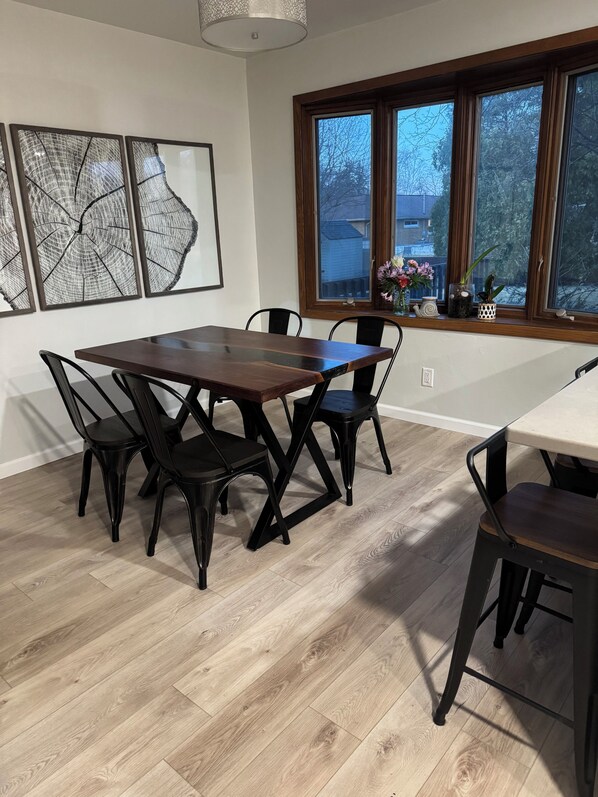 A dining area with a wooden table and chairs, a large window with a view of trees and houses, and three framed artworks on the wall. - Spacious 4-bedrm, 3 bath home in FDL, new kitchen, gym, WiFi, patio, 2 tv rooms (Fond du Lac)