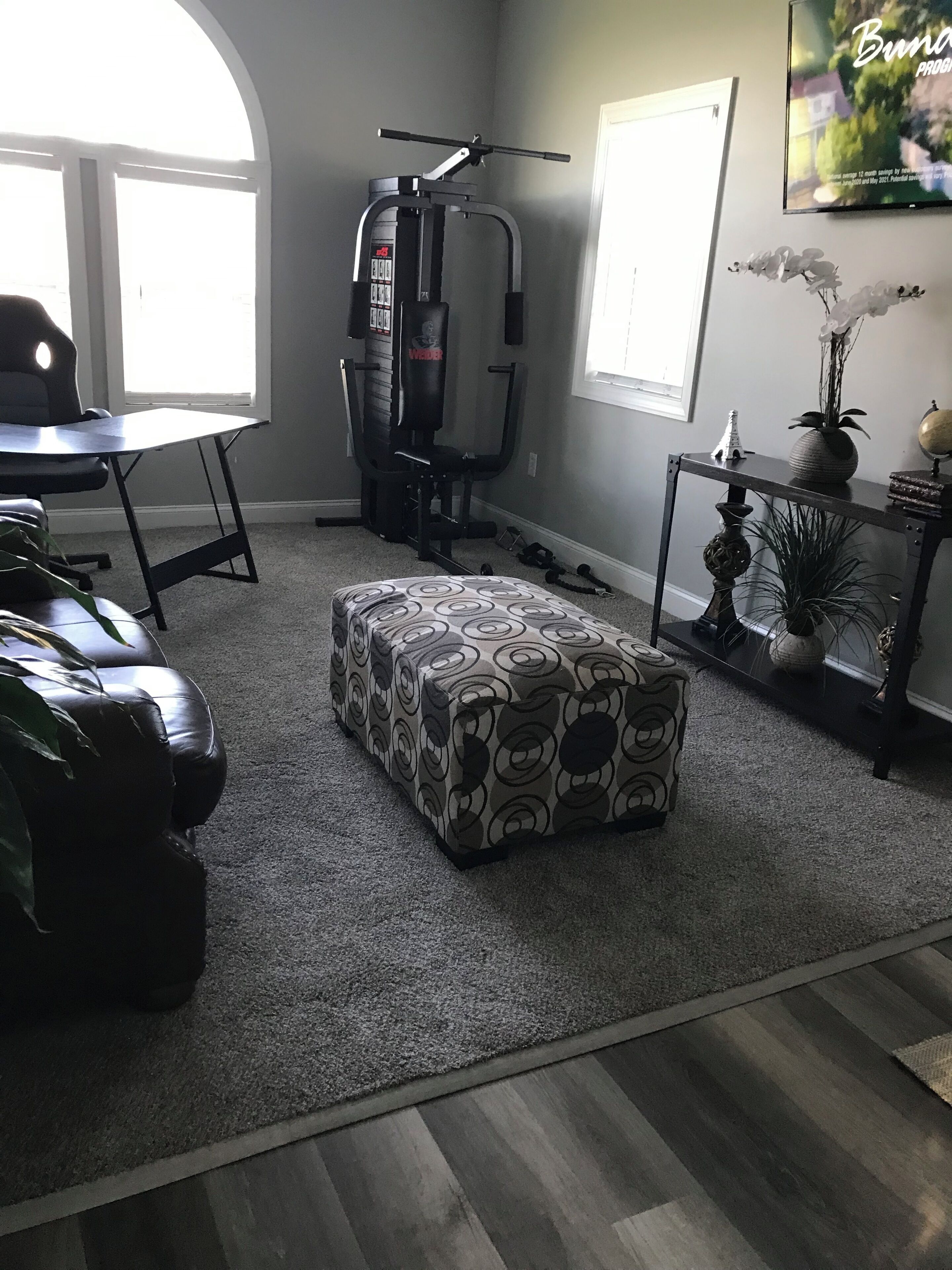 A living room with a grey carpet, a patterned ottoman, a leather couch, a desk, and a punching bag.