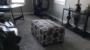 A living room with a grey carpet, a patterned ottoman, a leather couch, a desk, and a punching bag.