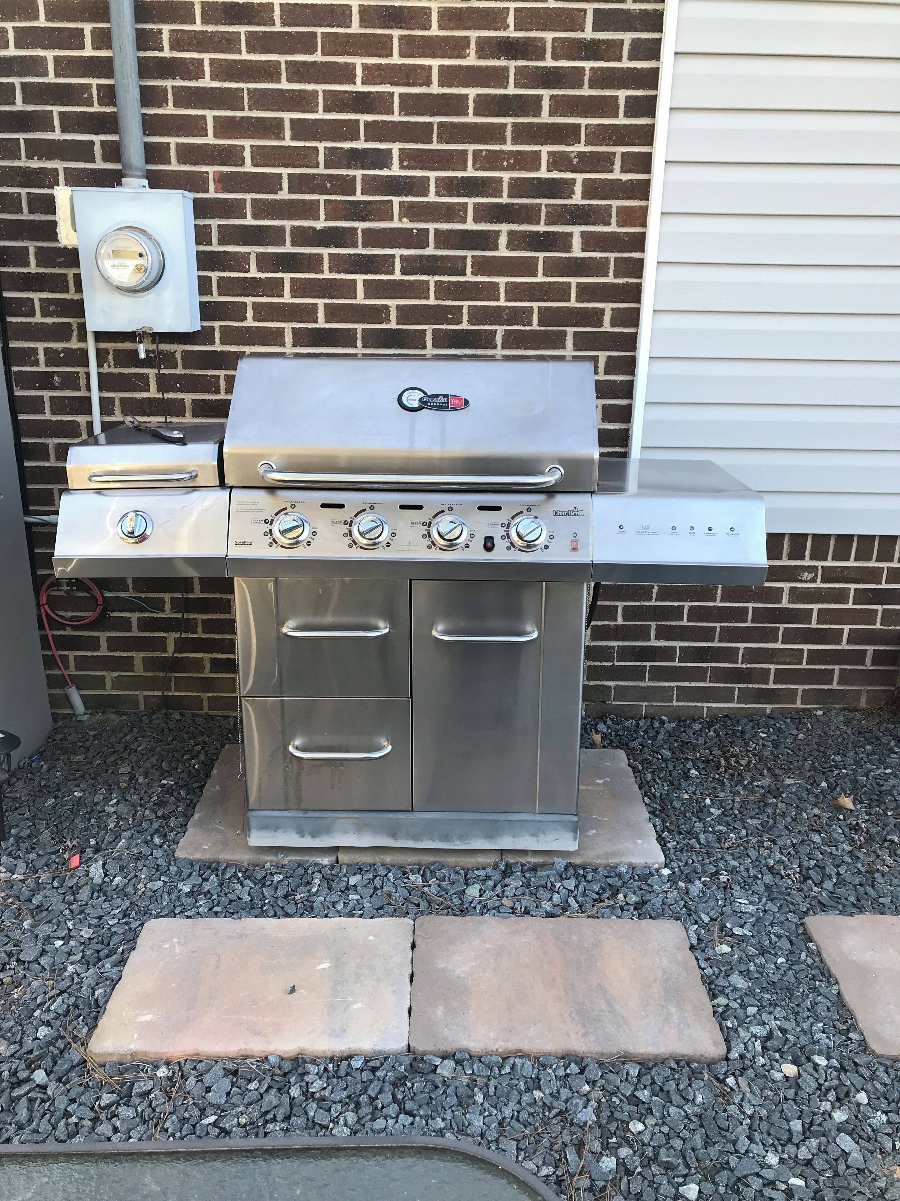 A stainless steel outdoor grill with four burners and a side burner, mounted on a brick wall with a utility box nearby.